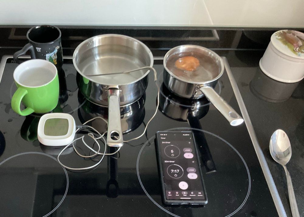 Egg cooking setup on a hob with two water-filled pots, a large spoon to transfer the egg, a timer, a temperature probe in one of the pots, and two mugs to transfer water.