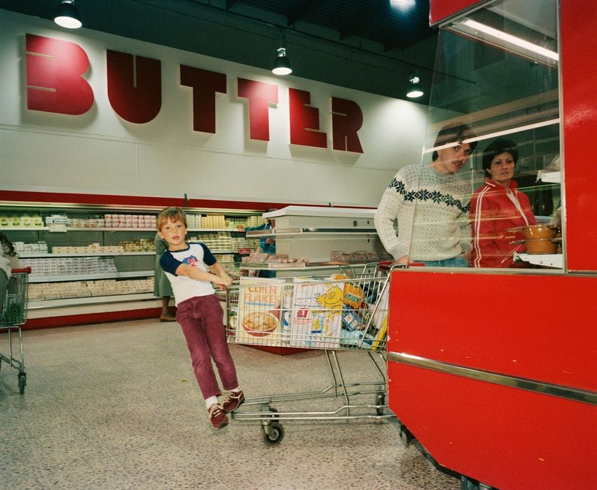 Photo a l’intérieur d’un lagasin Tesco. En arrière plan une gigantesque enseigne "Butter" au premier plan un morceau de tête de gondole de rayon frais rouge vif a travers laquelle on voit une femme en rouge aussi et un homme en pull blanx a motif tenir un caddie qui dépasse  de la gondole. Au bout du caddie, un enfant  s’agrippe et se penche a moitié dans le vide. Il a capté le photographe et le regarde. Son pantalon bordeau s’accorde avec le rayonnage en arrière plan.