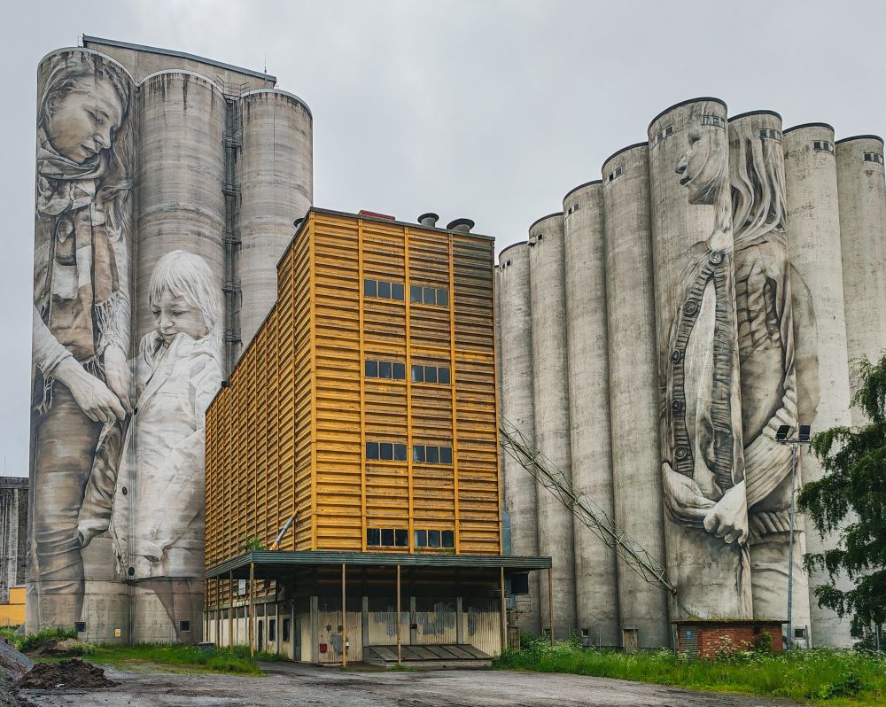 Viljasiiloihin maalattu muraali, joka kuvaa aikuista naista ja kahta pientä lasta. Siilojen välissä hylätyn näköinen rakennus.