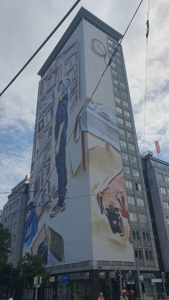 Der Ringturm in Wien ist mit einem Kunstwerk des Krakauer Künstler Marcin Maciejowski verhüllt. Man sieht einen Mann vor einem Bücherregal, einen Esstisch auf dem eine Tasse Kaffee steht und ein aufgeschlagenes Buch liegt und darunter einen liegenden Hund.