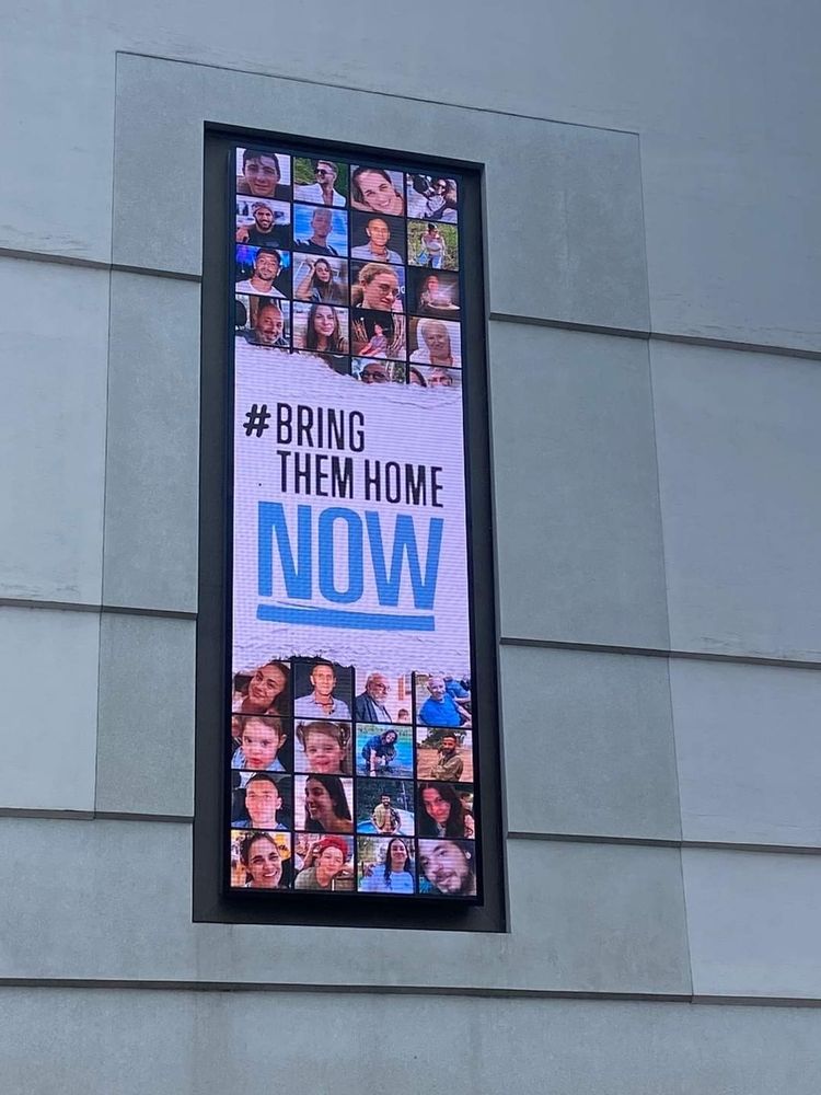 Die Medienfassade des Jüdischen Museums Frankfurt zeigt eine Grafik mit dem Schriftzug "BRING THEM HOME NOW". Im Hintergrund sieht man die Portraits der Verschleppten.