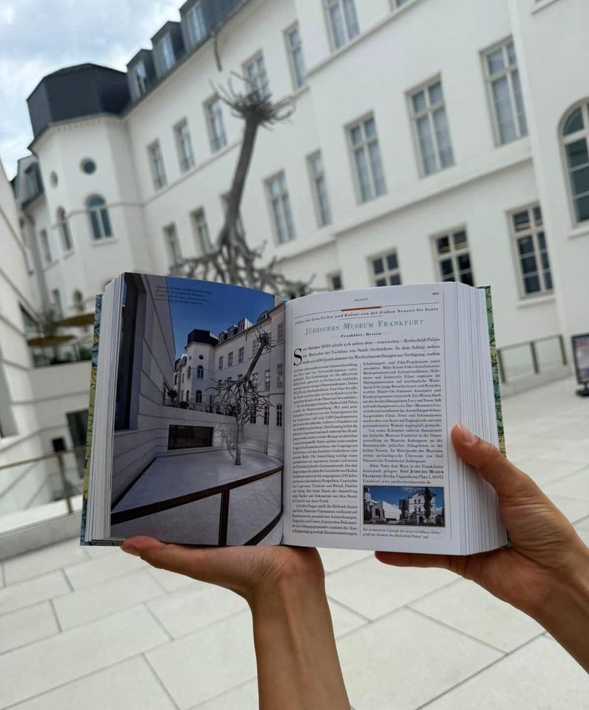 Vor dem Jüdischen Museum Frankfurt halten zwei Hände halten einen Reiseführer hoch: Darauf zu sehen aus der gleichen Perspektive ist ebenfalls das Jüdische Museum Frankfurt. 
