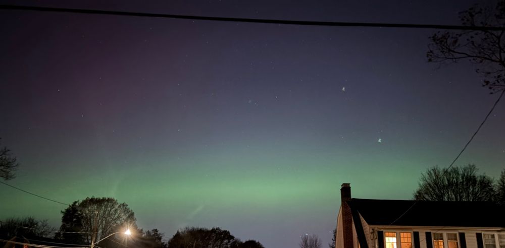 A solid green band of aurora in the sky, over the line of trees and the roof of a house.