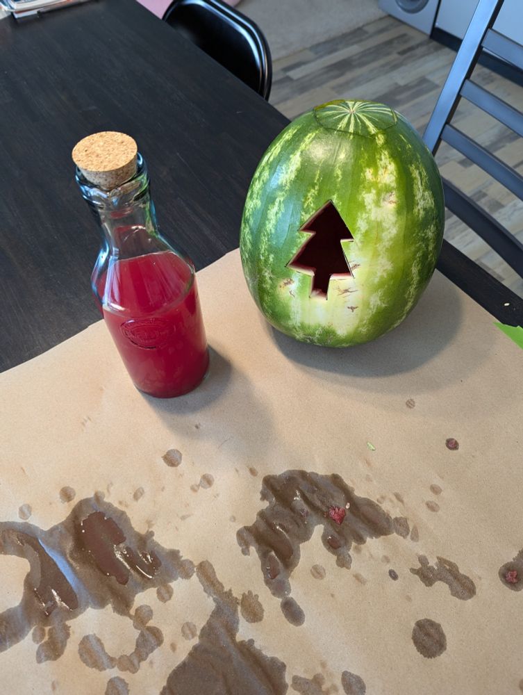 A pumpkin with a pine tree carved in it next to a full pitcher of watermelon juice.