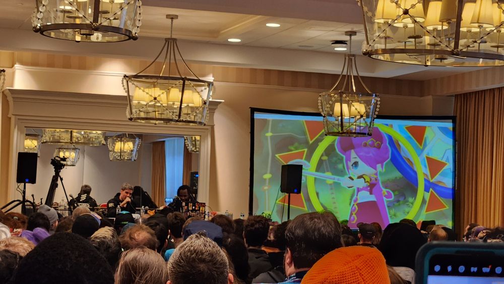 Super Eyepatch Wolf & WoolieVersus sitting together at their panel while the intro for Dokapon Kingdom Connect plays on the projector.