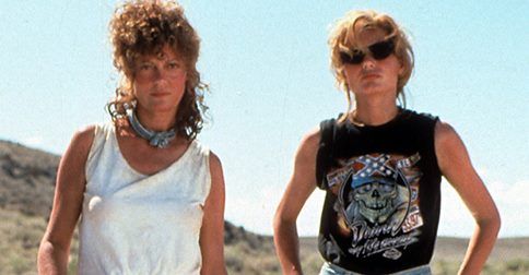 Susan Sarandon as Louise Sawyer and Geena Davis as Thelma Dickinson stand side by side in a desert setting from the movie Thelma & Louise. Louise, on the left, wears a sleeveless white top with a scarf tied around her neck and has curly hair. Thelma, on the right, wears dark sunglasses, a sleeveless black graphic T-shirt, and has tied back blonde hair