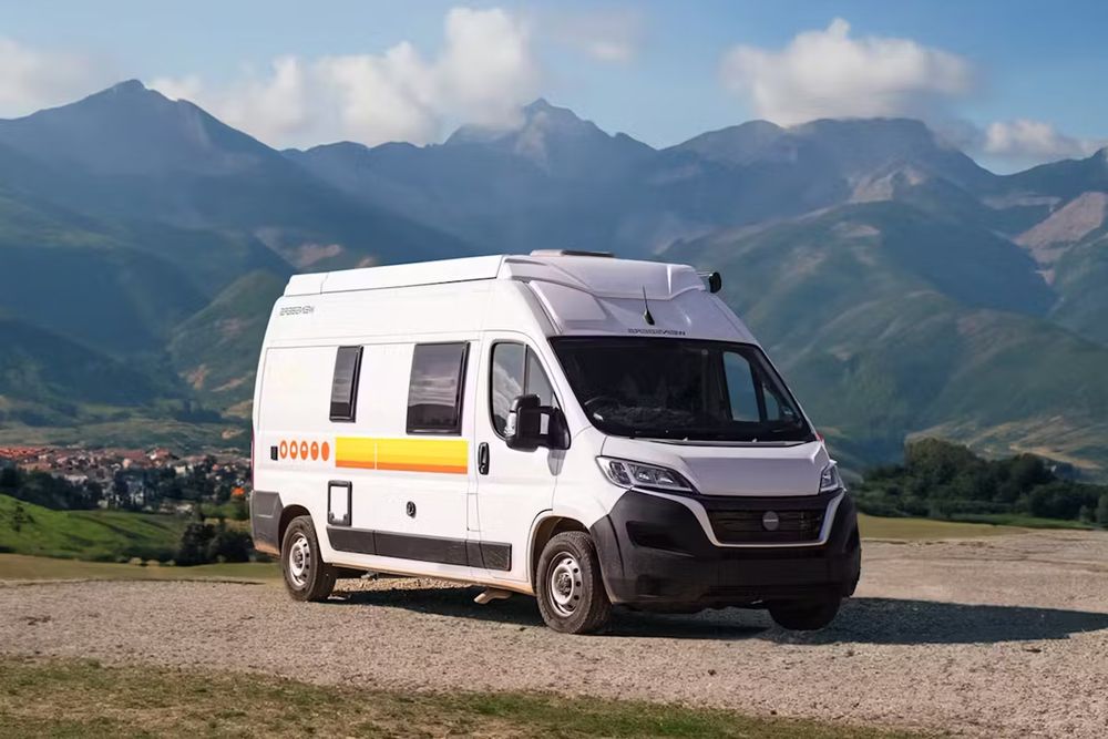 A white campervan with yellow and orange stripes parked with beautiful mountains in background. 