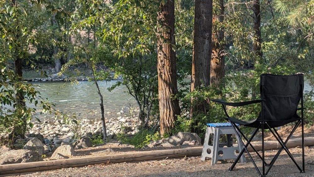 A riverside campsite with a camp chair and stool under tall trees next to a river.