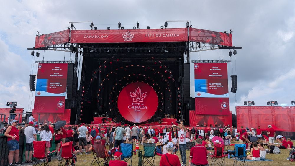 Large outdoor stage in an open field, draped in red with large screens on each side. Canadian symbols abound. People gathered in front are wearing red and white.