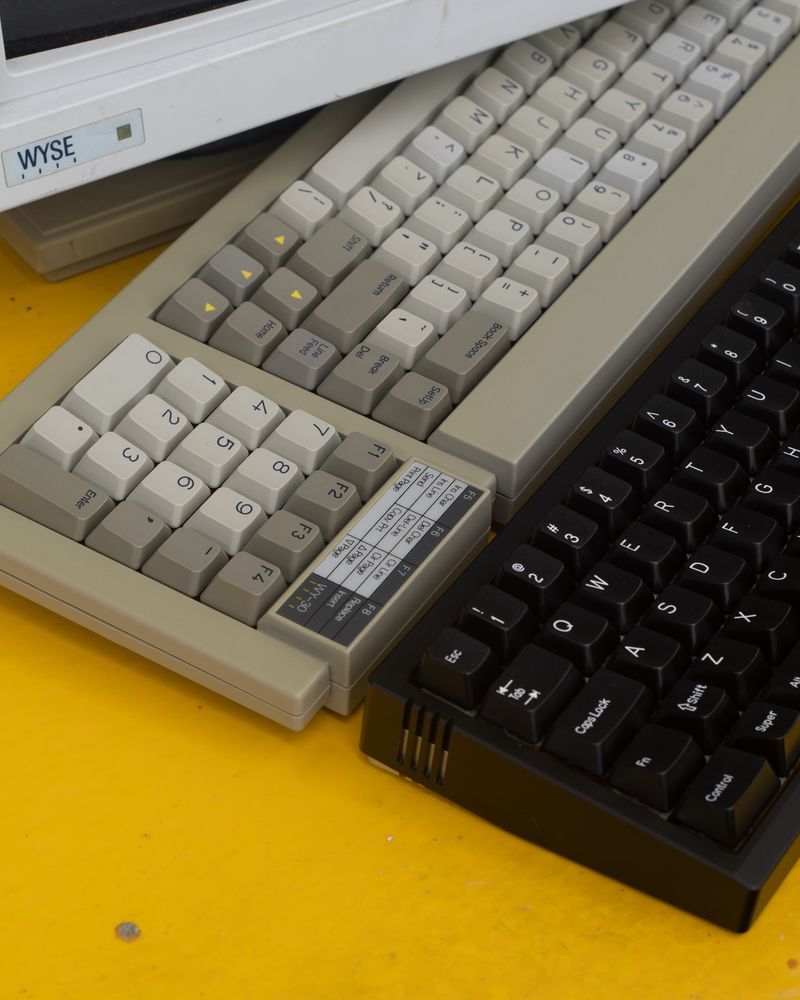 two mechanical keyboards, one beige, one black, on a yellow desk with the corner of a Wyse monitor above them.