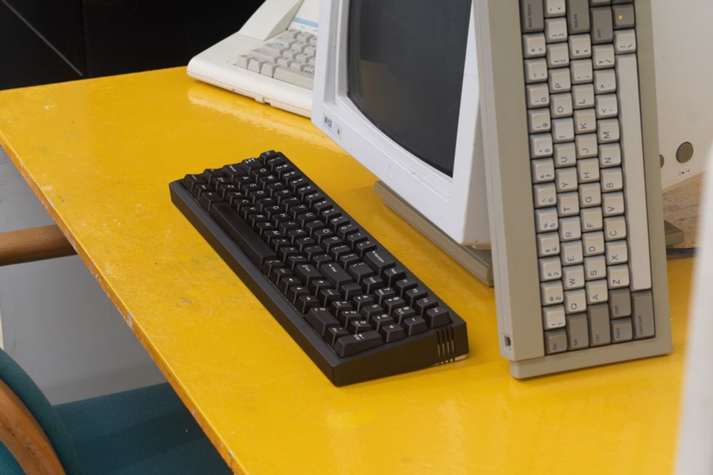 an ASCII-F keyboard in black sits in front of a Wyse monitor with a Wyse WY30 propped on the side of it. there is a typewriter in the background, and they all sit atop a bright yellow desk, with a teal cushioned chair to the left of it. 