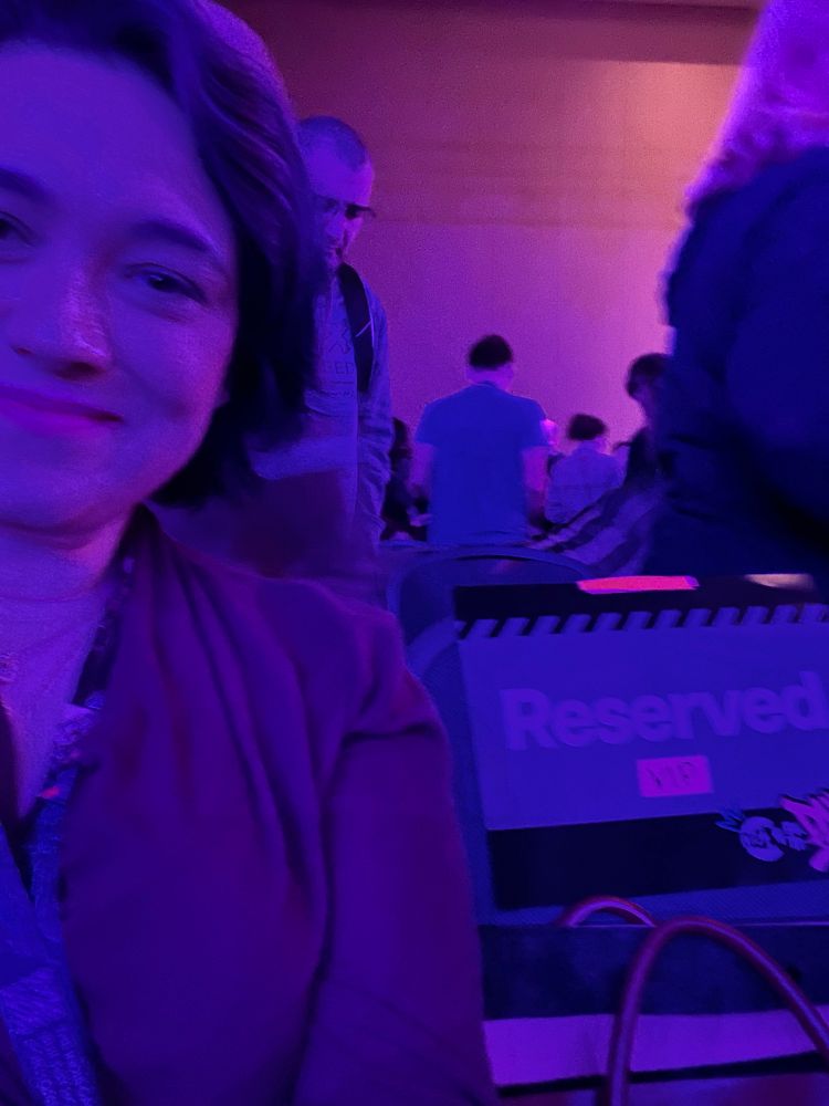 Emily, in blue and pink lighting, smiles next to a RESERVED seat like the one she is sitting in