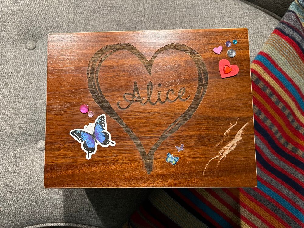A wooden box photographed from the top. Alice is carved inside the top within a large heart, and a variety of stickers, including a butterfly and hearts are stuck on top. A large gouge runs across one corner.