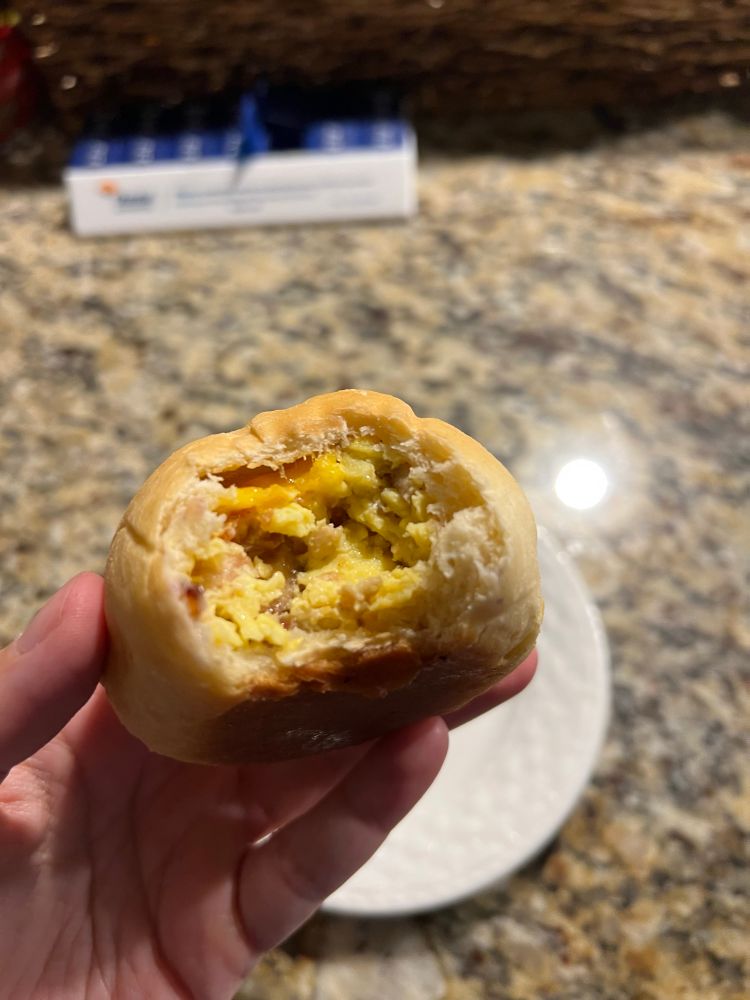 A kolache, a soft bread roll filled with egg, bacon, and cheese. A bite shows the interior.
