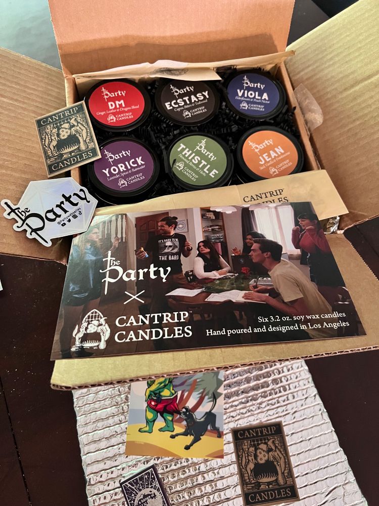 Insulated cardboard box that has a smaller cardboard box inside it. That box includes six candle tins, each with a different colored label, and the name of one of the characters from the web series, the party. Cards & stickers from the show & from the candlemaker Cantrip Candles are scattered.