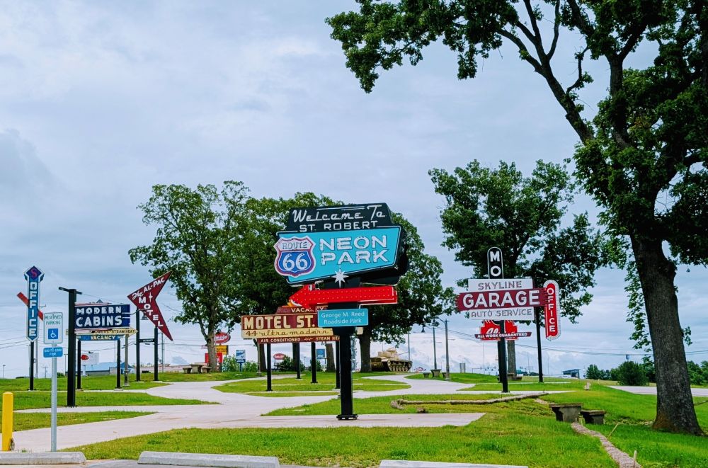 Several neon signs stand in a roadside park. Welcome to St Robert Route 66 Neon Park front and center. Various signs advertising motels and garages.

