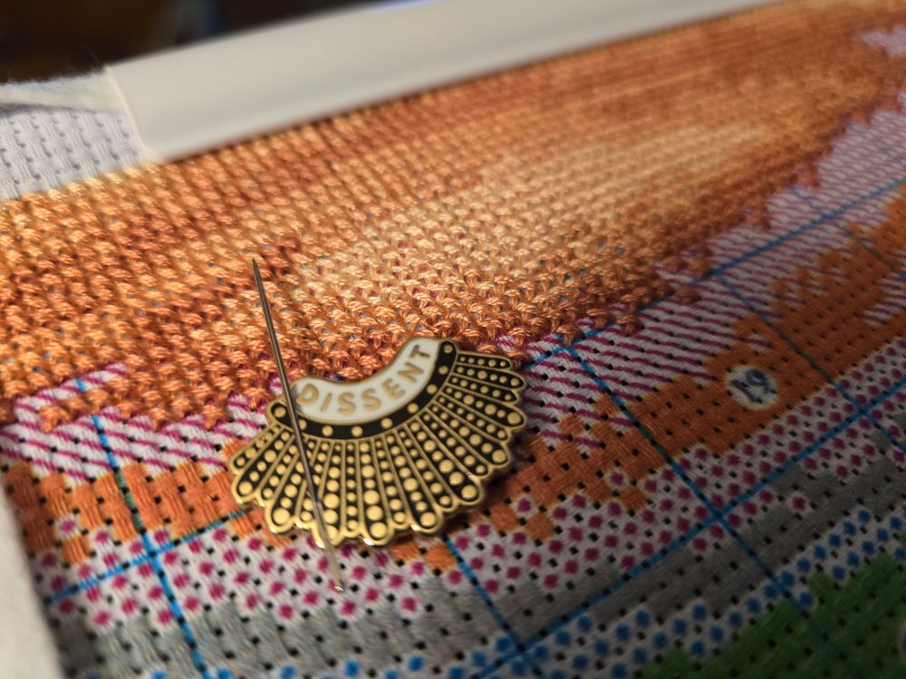 Stamped #crossstitch partially complete with a bunch of orange colors which is going to be the sky. A needle minder of RBG's "dissent" collar with my needle attached in view. #stitchsky #craftsky
