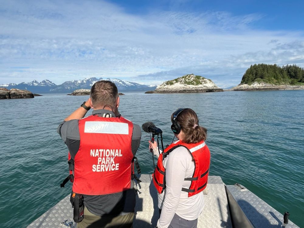 Two people on a boat, recording footage of the mountains beyond.