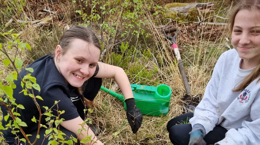 Two smiling school pupils crouched on the scrubby ground, planting a sapling