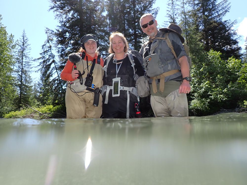 Megan Klaar smiling with the podcast producers, standing in a river with trees behind.