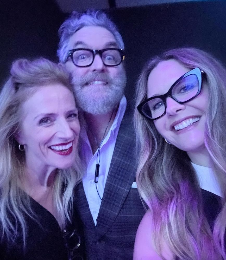 Three smiling friends sharing a happy moment backstage for the camera. A man and woman are wearing glasses. One woman is wearing bright red lipstick. 