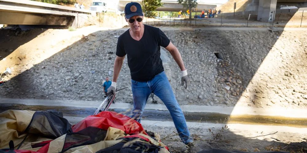 Publicity photo from August 2024. Newsom, in a cap, sunglasses, jeans, and black T-shirt, holds what appears to be a tent. 