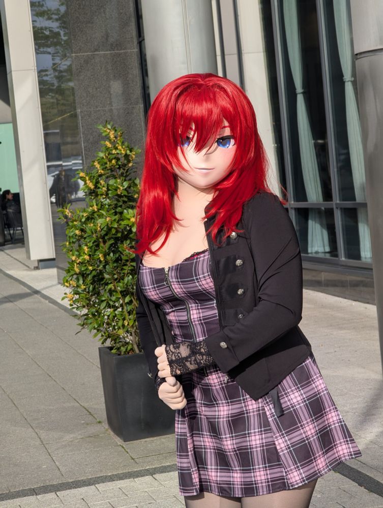 Scarlett posing in front of a hotel, wearing a pink-and-black plaid minidress, lace gloves, tights and a black jacket.