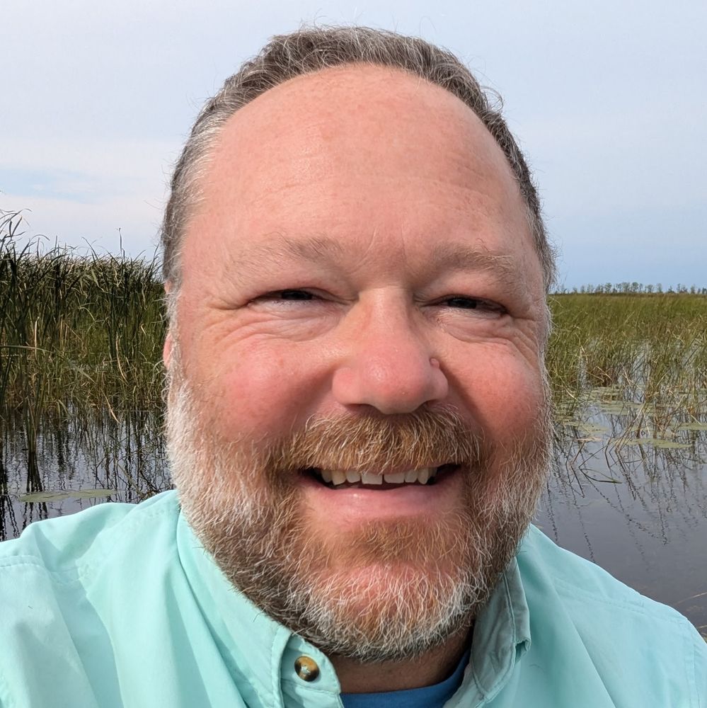 Headshot (yikes) of male with graying beard, it was once all reddish, and a receding hairline but he's smiling because he is in a Great Lakes coastal wetland!