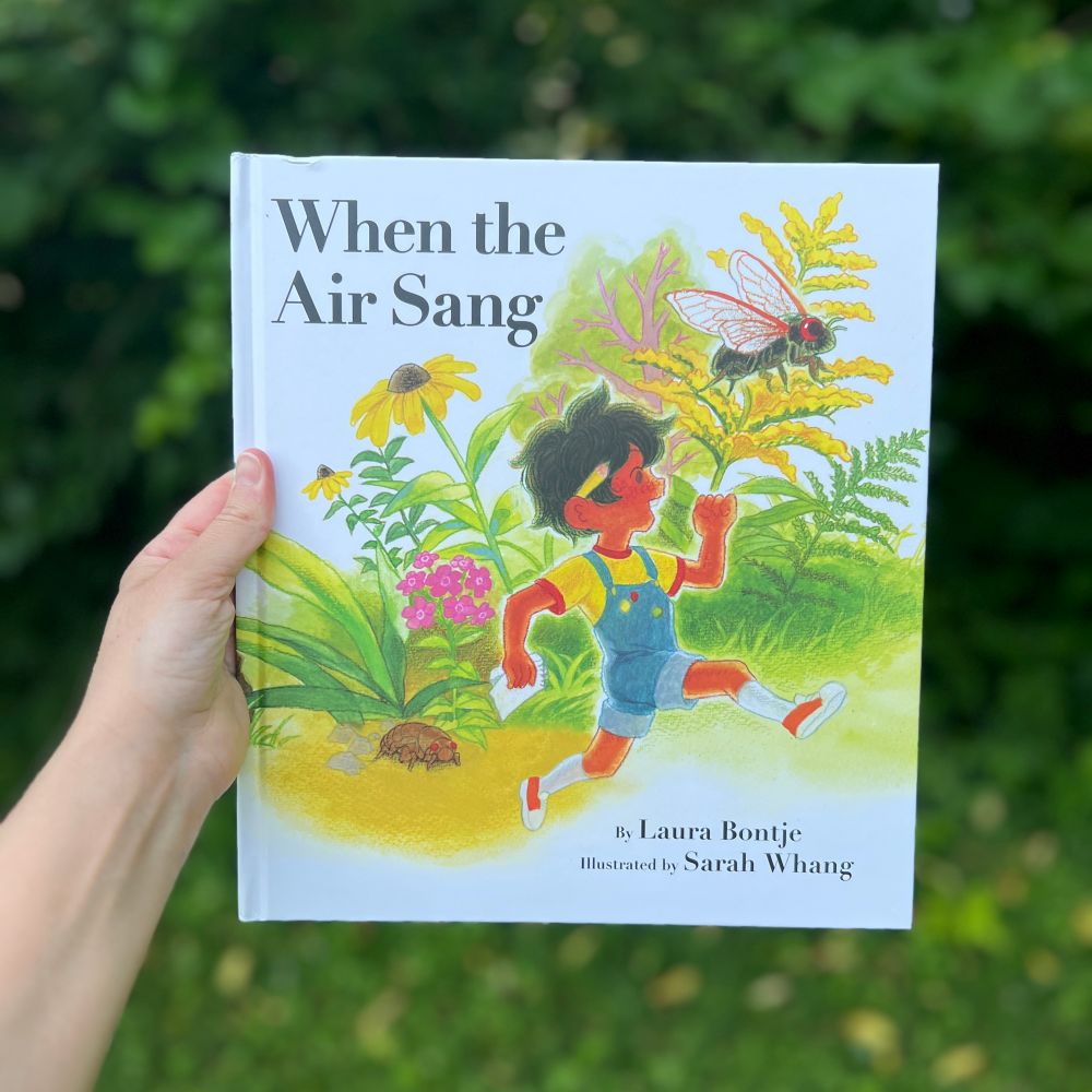 1. My hand holds the book in front of trees. The white cover shows a brown skinned, black haired child running through an oversized garden, a cicada on a yellow flower.