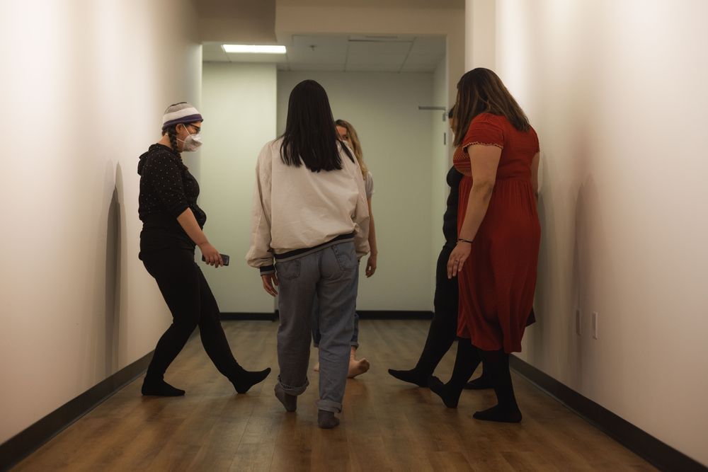 Jo leads a group of dancers in a circle. They are all sticking out a flexed foot into the center of the circle. 