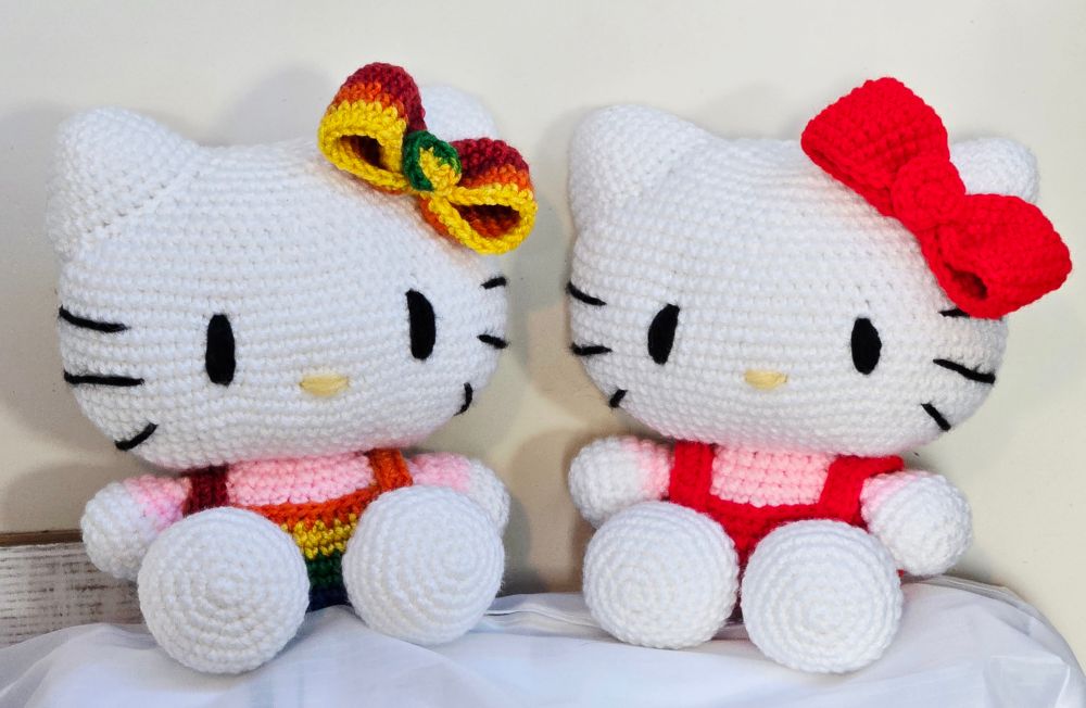 Two crochet plush toys of Hello Kitty. One has red overalls and bow with a pink shirt, the other has rainbow overalls and bow with a pink shirt.