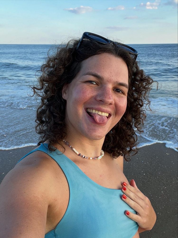 me at the beach this summer in a light blue bikini top smiling and sticking my tongue out as i stand in front of the ocean