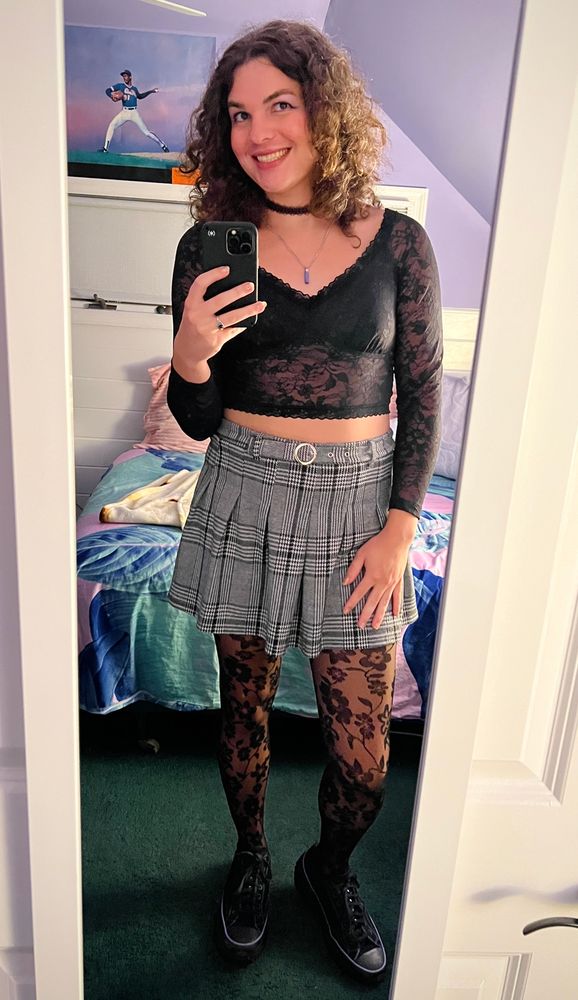 mirror selfie of me smiling, wearing a black lace choker, silver necklace with purple pendant, black flower lace v-neck long-sleeve crop top, black/white/gray houndstooth patterned pleated skirt with a little belt built in, black flower lace tights, and black and purple platform converse sneakers