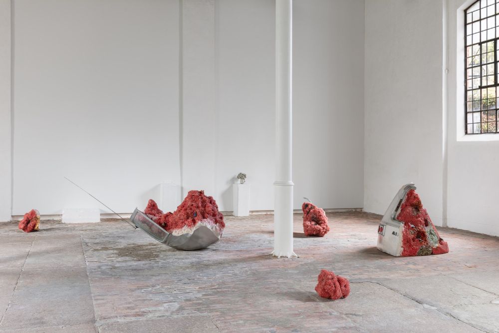 Image shows a large white gallery space with tall white support beam a little off center. On the floor, the artist installed five irregular fragments that resemble fragments of a giant, rotting, smashed watermelon. The rind of the watermelon is made out fragments of the shell of an Airstream camping trailer. The pink pulp of the fruit is made out of semi precious stones with a few rusty cast-iron flies crawling over the mouldering flesh. Imagine what an AirStream trailer watermelon would look like before it was smashed. The shape resembles a watermelon.