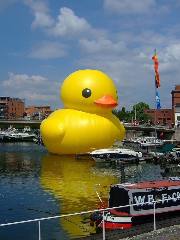 The photograph shows a four-story high inflatable rubber duck floating in a harbor full of boats by a seaside town.