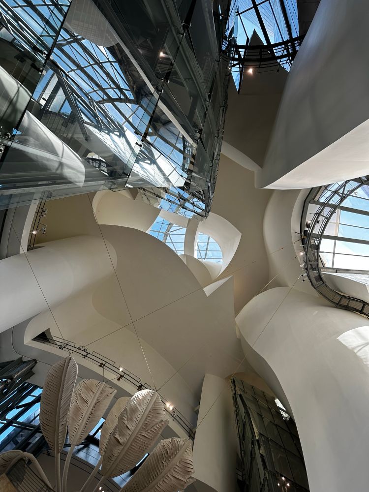 Photo taken inside the Guggenheim Museum Bilbao, looking up towards the ceiling. A jumble of pillars cans walkways is visible, intermingled with windows through which sunshine and blue sky may be seen.