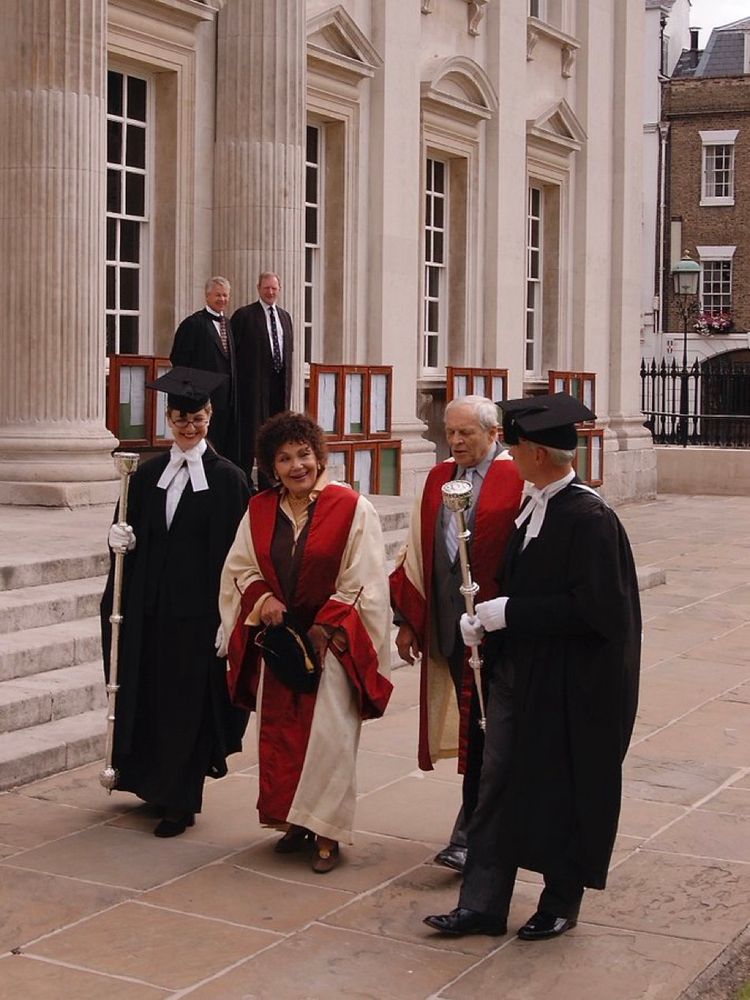 Photograph of four people in academical dress walking in front of a large stone building. Two are dressed in black gowns and are carrying silver maces. The other two (Dame Cleo Laine and John Dankworth) are dressed in colourful gowns of cream damask silk with cherry-coloured silk at the end of the sleeves and on the facings. Two men in black gowns are seen in the background, standing on the steps of the building, watching the group as they walk by.