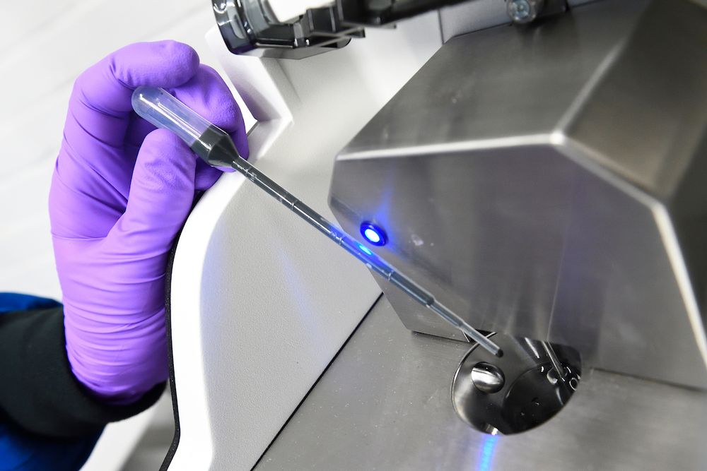 Gloved hand inserts a sample into a device emitting a blue light, likely for particle size or stress testing in cathode materials.