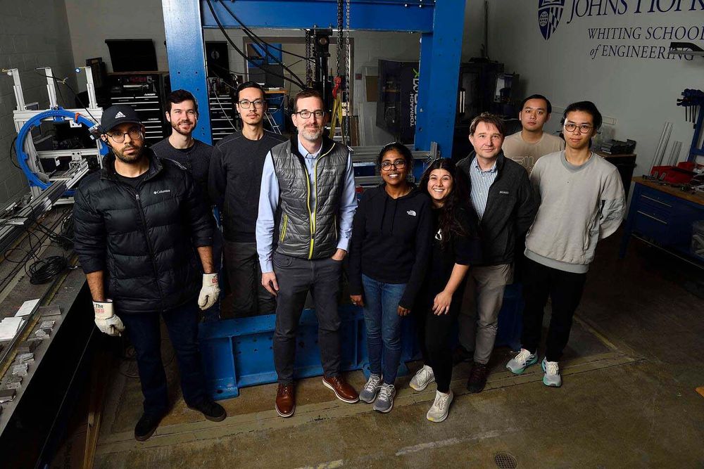 Ben Schafer and 8 members of his lab pose next to industrial testing equipment 