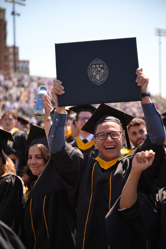 Cheerful graduate holding up a diploma folder with the Johns Hopkins seal, surrounded by classmates in caps and gowns.