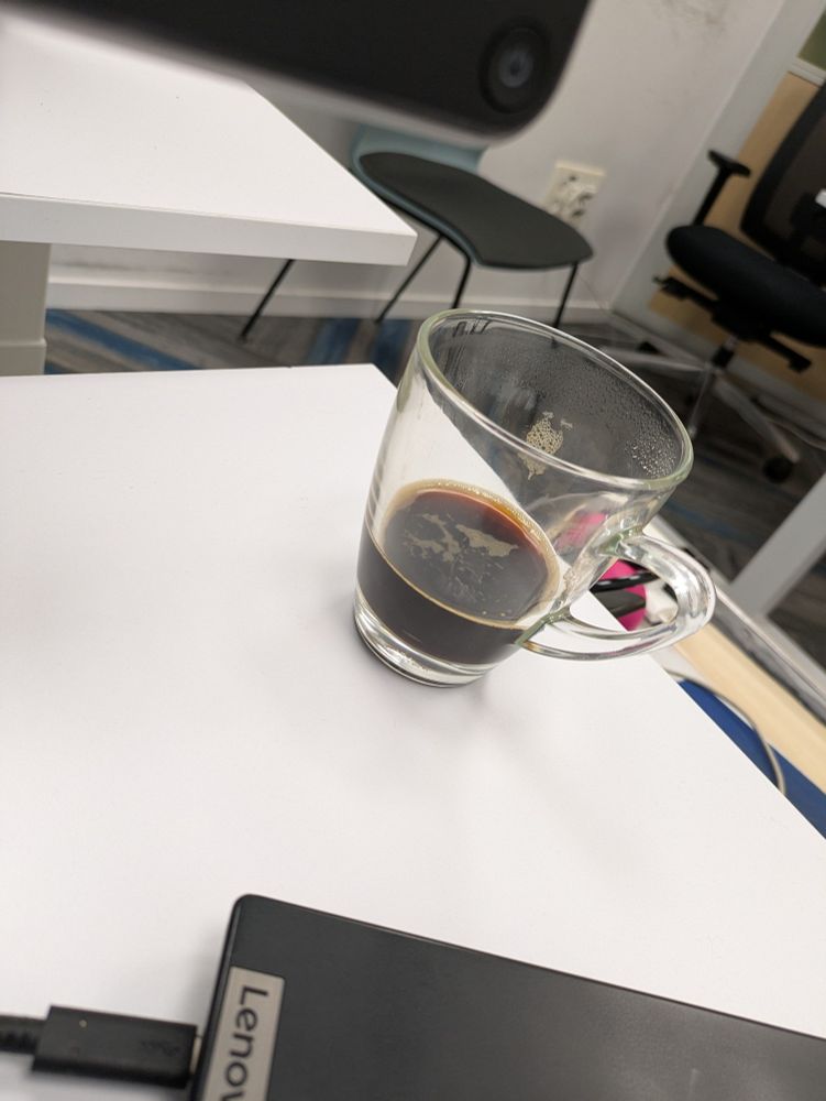 כוס קפה (גדולה) מלאה כדי שליש מתכולתה, אחרי שמחצית מהמשקה השחור נלגמה. סימני קצף חום כהה על צדה הפנימי של הכוס.
הכוס מונחת על שולחן לבן, סמוך לה לפטופ שחור עם כיתוב Leno. ברקע ציוד משרדי על רקע אפור כללי דהוי