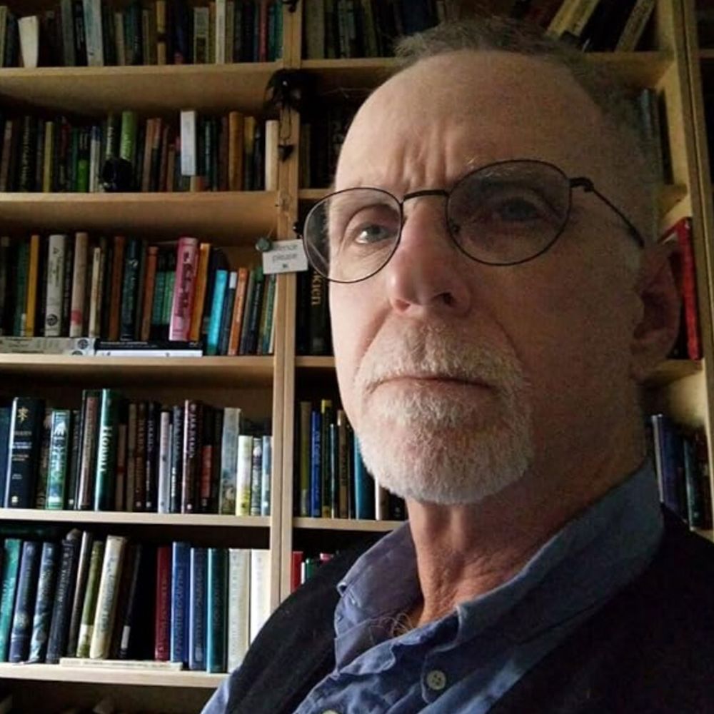 A man, Thomas P. Hillman, with a beard and glasses stands before a bookcase, which is lined with numerous books of different sizes and colors.