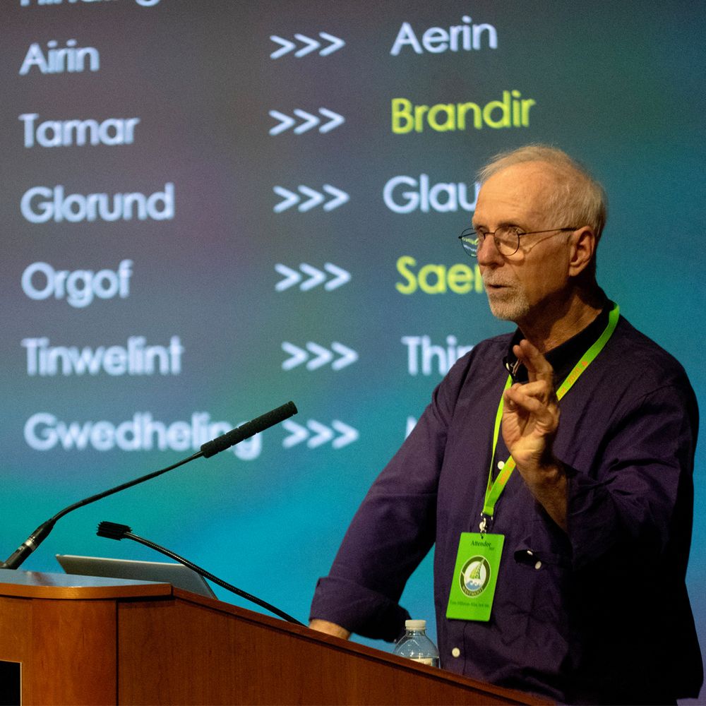 A presenter, Tom Hillman, stands confidently at a podium, addressing an audience with a focused expression.