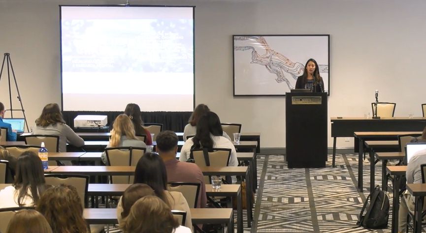 Audience members sit in a hotel conference room facing Dr. Agtarap, who stands at a podium at the front. Next to her is a presentation screen presenting the title of her talk: "Associations between posttraumatic growth and depreciation, environmental limitations, and psychosocial outcomes following moderate-severe TBI: Findings from a mixed-methods pilot study".