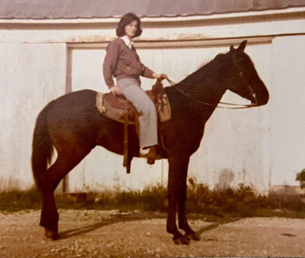 Photo of a woman in her 20s with shoulder length hair in a jacket and trousers seated on a dark brown horse circa 1975