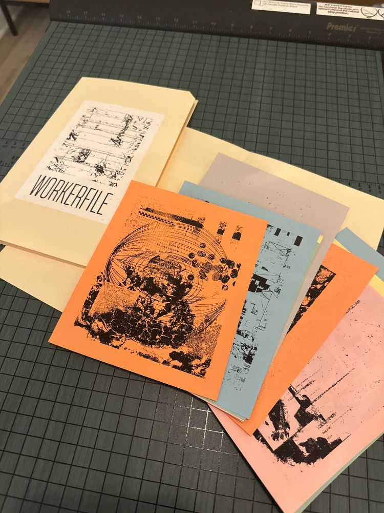 On top of a large paper cutter are two manila folders. One of the folders is closed, a 4x6 inch sticker with abstract black and white art and the word "WORKERFILE" under it. The other folder is open, showing other black and white abstract works, printed on colored pieces of paper at various sizes.