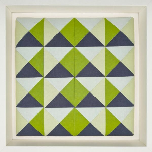 A 12“ quilt block with 12 hourglass blocks in grays and greens, mounted on canvas stretchers and framed by a white shadow box.