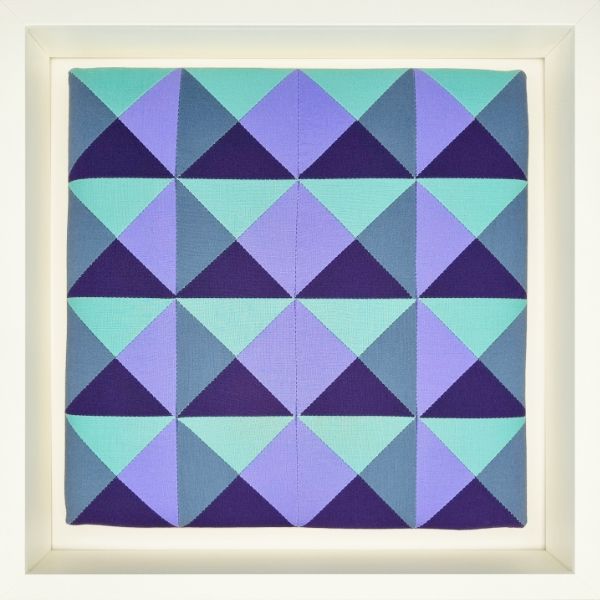 A 12“ quiltblock with 16 hourglass blocks in purples and greens, mounted on canvas stretchers and framed with a white shadow box.