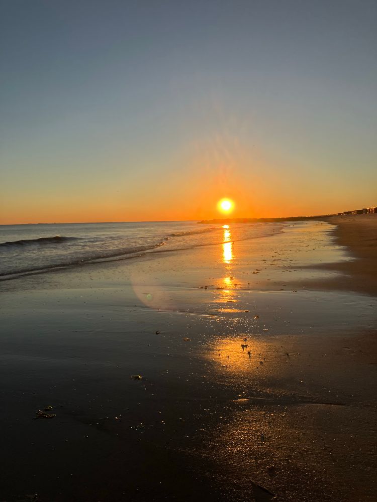 An orange sun dips toward the horizon and reflects on the wet, flat sand