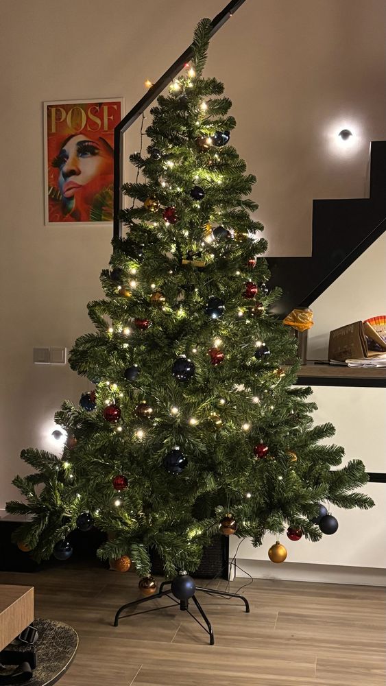 A Christmas Tree in a flat against a white background. It is decorated with white fairy lights and baubles of various colours: copper, navy, and red. It’s very festive!